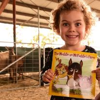 young girl holding a children's book about a farm in front of cows in a pen